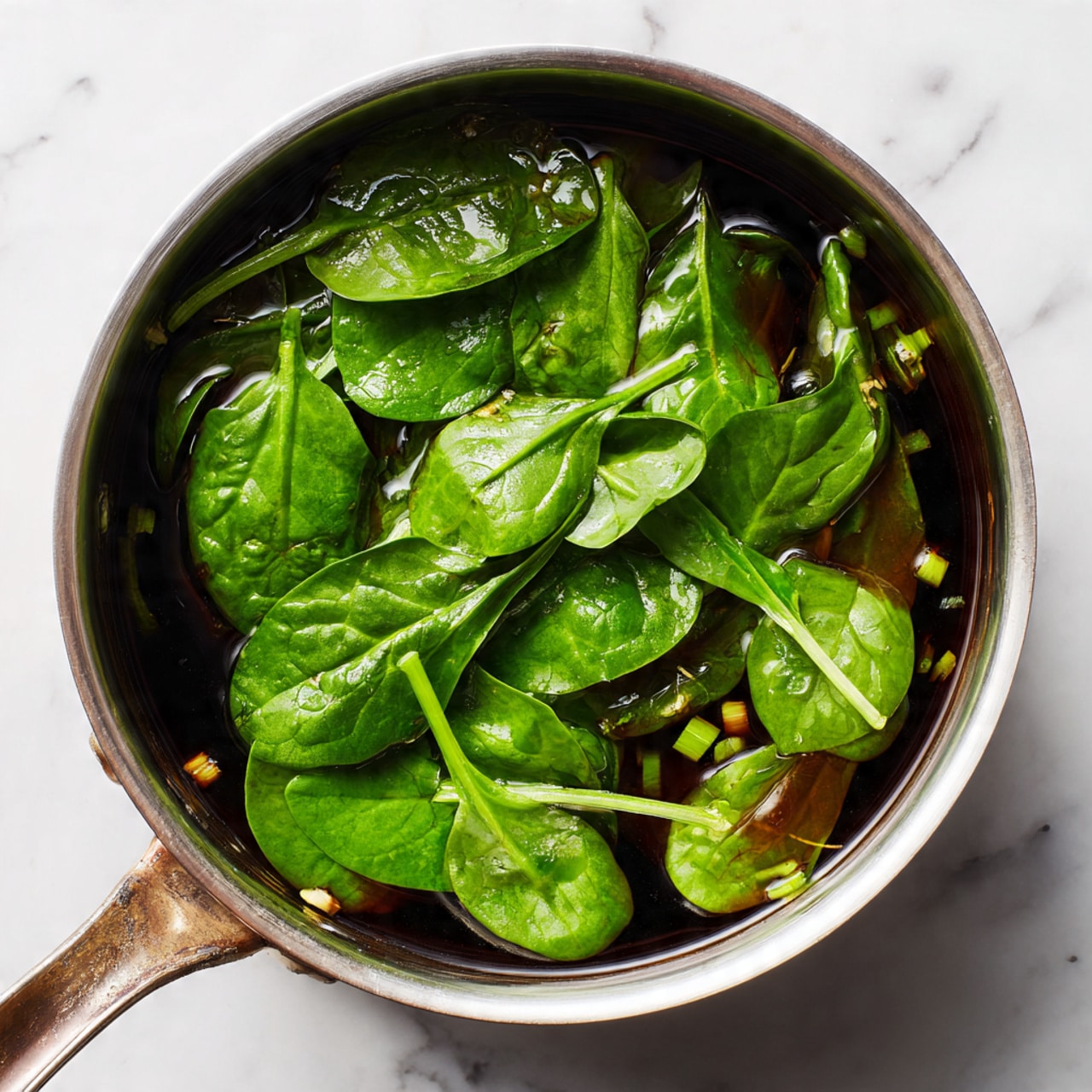 A silver metal pot sits on a white marbled surface, filled with several fresh green spinach leaves layered on top. Below the spinach is a dark brown broth that looks clear and glossy, showing small green onion pieces floating within. The pot handle is visible on the left side, catching a bit of light. The spinach leaves look fresh and slightly shiny, covering most of the broth underneath. Photo taken with an iphone --ar 4:5 --v 7