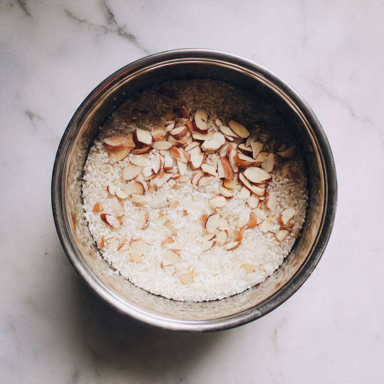 A silver metal pot sits on a white marbled surface, containing a single layer mix of small white rice grains and light brown sliced almonds spread evenly across the bottom. The pot's smooth inner wall reflects some light, and the rice and nuts rest quietly inside without any visible liquid. Photo taken with an iphone --ar 4:5 --v 7