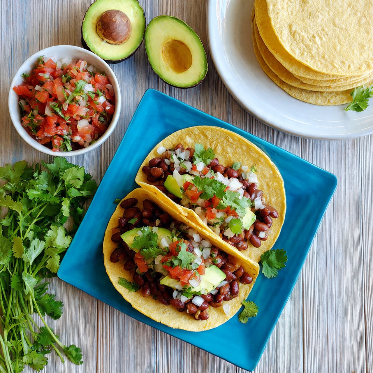 The image shows two soft yellow corn tortillas on a bright blue square plate, each tortilla layered with brown beans at the bottom, topped with white chopped onions, red diced tomatoes, and a light green avocado slice angled on one side. Fresh green cilantro leaves are scattered on top and around the tacos, adding color contrast. Beside the plate, there is a bunch of fresh cilantro on a white marbled surface, and two avocado halves with a rich green inside and dark brown seed are placed near the top left. To the right, there is a stack of pale yellow corn tortillas on a white plate and a small white bowl filled with the same onion and tomato mix used on the tacos. The background consists of light wooden slats. photo taken with an iphone --ar 4:5 --v 7