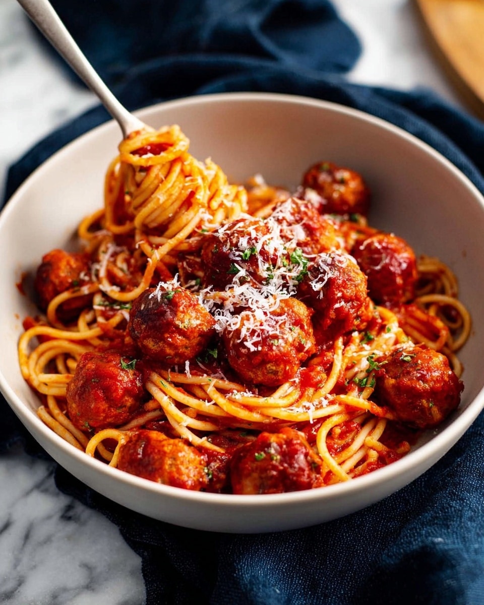 A white bowl filled with a base layer of spaghetti noodles coated in bright red tomato sauce, topped with many small, round meatballs covered in the same sauce. The meatballs have a rich, slightly glossy texture and are scattered evenly over the pasta. A generous sprinkling of finely grated white cheese covers the meatballs, with small bits of green herbs for contrast. A silver fork rests inside the bowl, twisting a few strands of spaghetti around its tines. The bowl sits on a dark blue cloth, placed against a white marbled background. Photo taken with an iphone --ar 4:5 --v 7