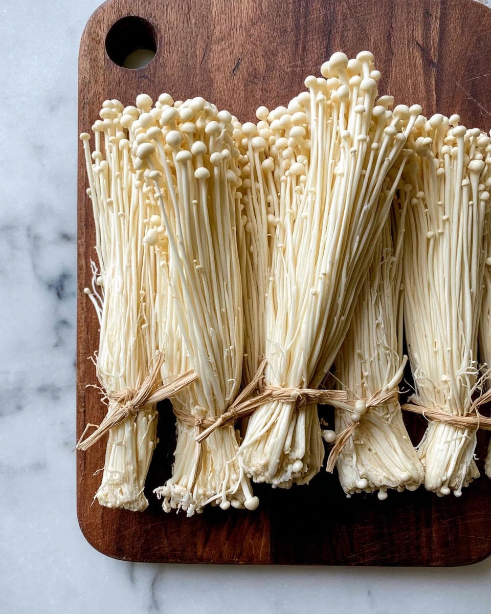 The image shows several bundles of enoki mushrooms with long, thin white stems and small round caps. Each bundle is tied together near the base with a natural string, and the mushrooms have a smooth, slightly shiny texture. These bundles are placed closely together on a brown cutting board that has a round hole in the top left corner. The cutting board is set on a white marbled surface, which adds a clean and bright contrast to the pale yellow-white mushrooms and the dark board. photo taken with an iphone --ar 4:5 --v 7