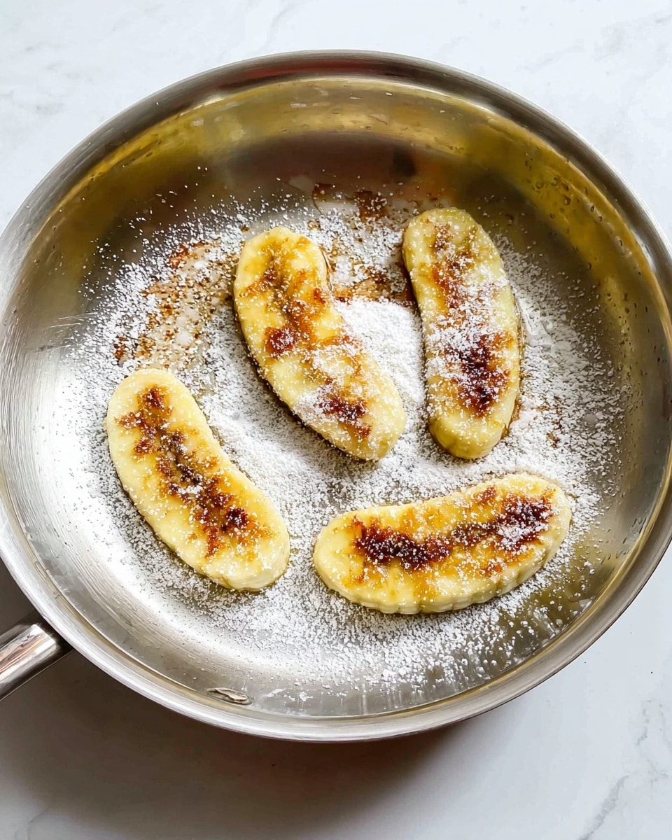 In a shiny silver pan, there are four slices of cooked banana laid flat in a circular shape. Each banana slice is golden yellow with some brown caramelized spots on top. A light layer of white sugar is sprinkled over the banana slices, giving a powdered look that contrasts with the warm banana color. The pan and the banana slices are set on a white marbled surface. photo taken with an iphone --ar 4:5 --v 7