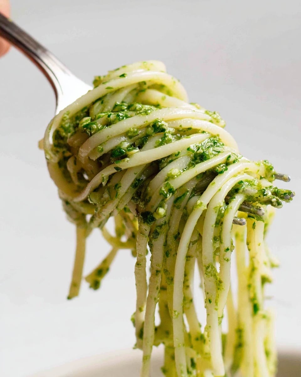 A clear glass bowl sits on a white marbled surface, filled with a thick, coarse green pesto sauce that has a slightly oily shine. The sauce texture is uneven, showing small bits of herbs and nuts. Partly immersed in the bowl is a stainless steel hand blender, blending the green mixture. The edges of the bowl have some pesto splatters. photo taken with an iphone --ar 4:5 --v 7