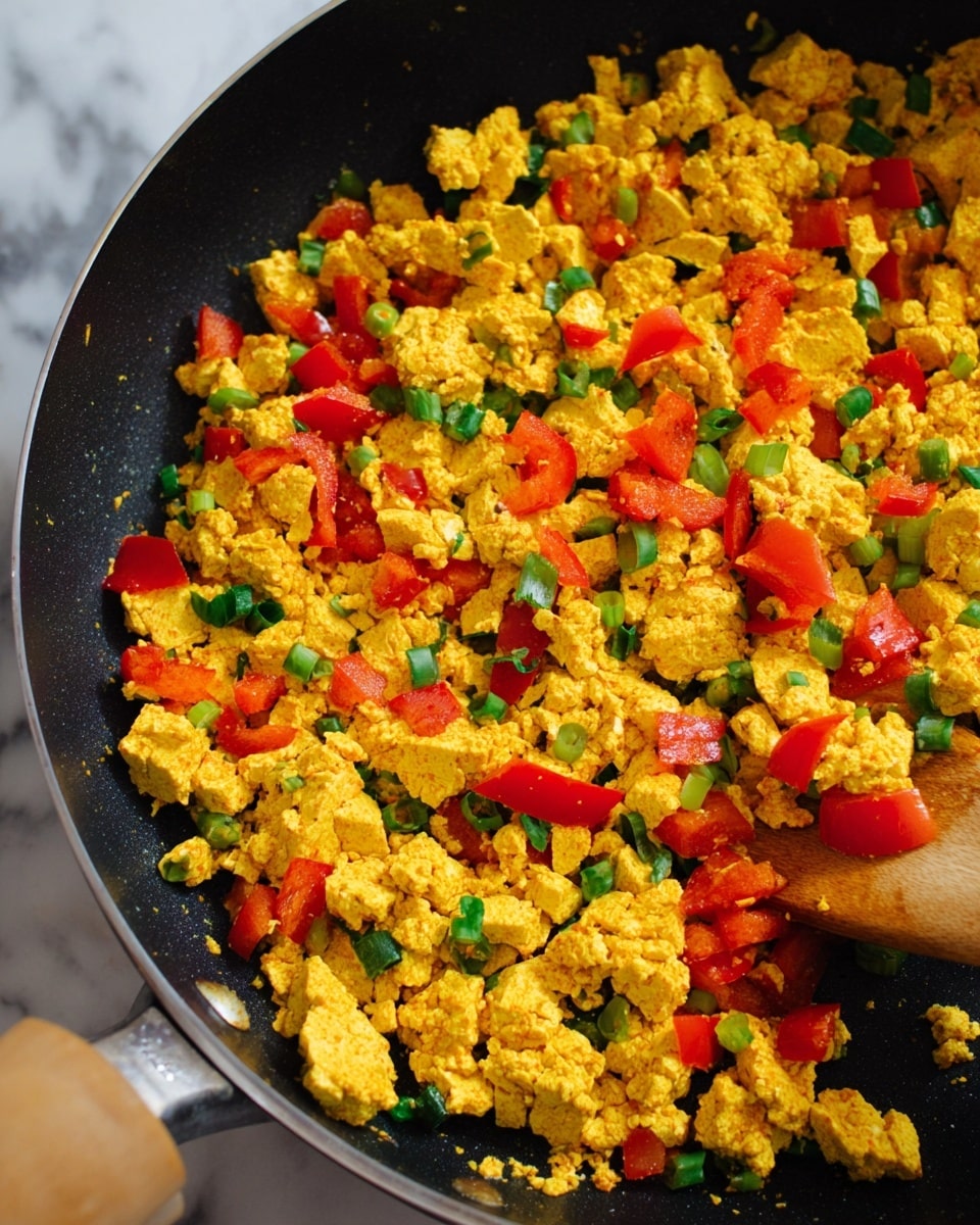 A close-up view of a black frying pan filled with bright yellow scrambled tofu pieces mixed with chopped red bell peppers and small green onion slices, giving a mix of warm yellow, vibrant red, and fresh green colors throughout. The tofu pieces have a soft, crumbly texture, while the vegetables add a slightly shiny and fresh look. On the bottom left corner, a wooden spatula is slightly visible, moving the tofu and vegetables inside the pan. The background shows a white marbled surface. Photo taken with an iphone --ar 4:5 --v 7