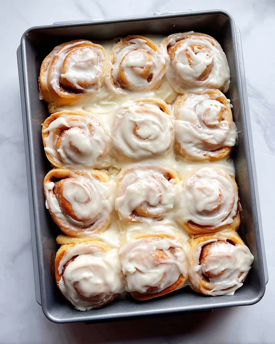 A metal baking tray holds twelve cinnamon rolls arranged in a tight 3x4 grid. Each fluffy roll is light brown with visible spiral layers of dough and cinnamon filling. A thick layer of white creamy icing is generously spread on top, dripping down the sides and filling spaces between the rolls. The tray is placed on a white marbled surface, giving a clean and bright background. photo taken with an iphone --ar 4:5 --v 7