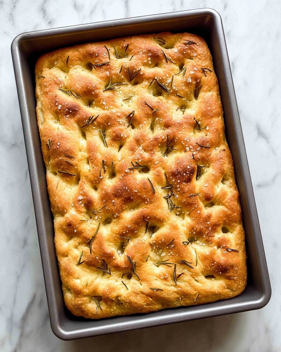 The image shows three square pieces of focaccia bread stacked on a white plate. Each piece has a light golden brown top with a slightly rough texture, and the inside is airy with visible holes. The top layer is sprinkled with coarse salt and small dark green herb needles, likely rosemary. The bread's edges are slightly more browned, adding a toasted contrast. The plate rests on a white marbled surface with a subtle gray vein pattern, and the background is similarly white marble. photo taken with an iphone --ar 4:5 --v 7