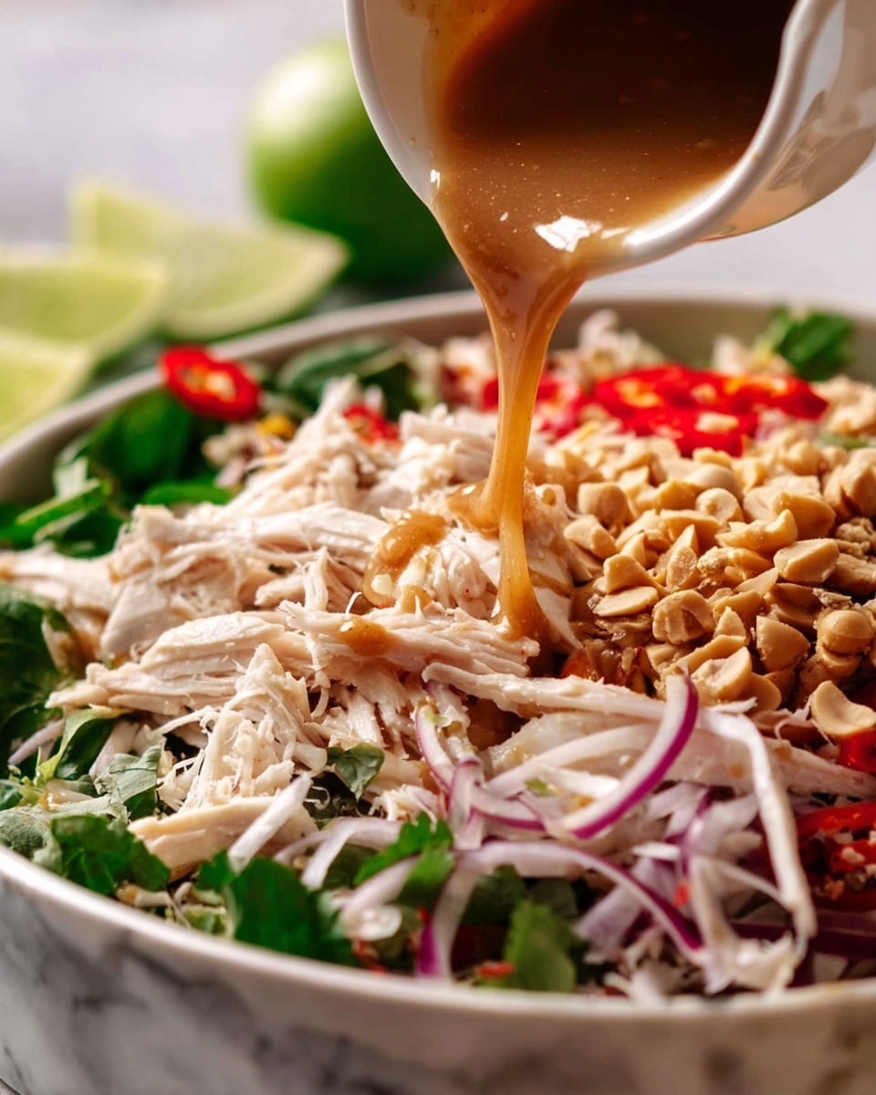 The image shows a close-up of a salad with several layers on a white marbled surface. The bottom layer is made up of shredded white chicken pieces mixed with green leafy herbs, thin red chili slices, and thin purple onion rings. On the right side of the salad, there is a generous layer of chopped peanuts. Above, a light brown sauce is being poured from a small white sauce pot, partially covering the chicken and nuts. In the background, green lime wedges add a fresh touch to the scene. The photo taken with an iphone --ar 4:5 --v 7