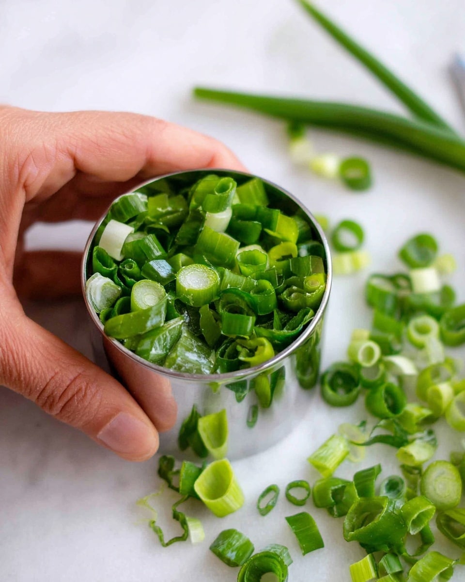 A woman's hand holds a silver round ring mold filled with bright green sliced green onions. Around the mold, more green onions pieces are scattered on a white marbled surface. The green onions have a fresh, glossy texture with different shades of green from light to dark. The silver ring mold reflects the green colors and the surroundings clearly. Photo taken with an iphone --ar 4:5 --v 7
