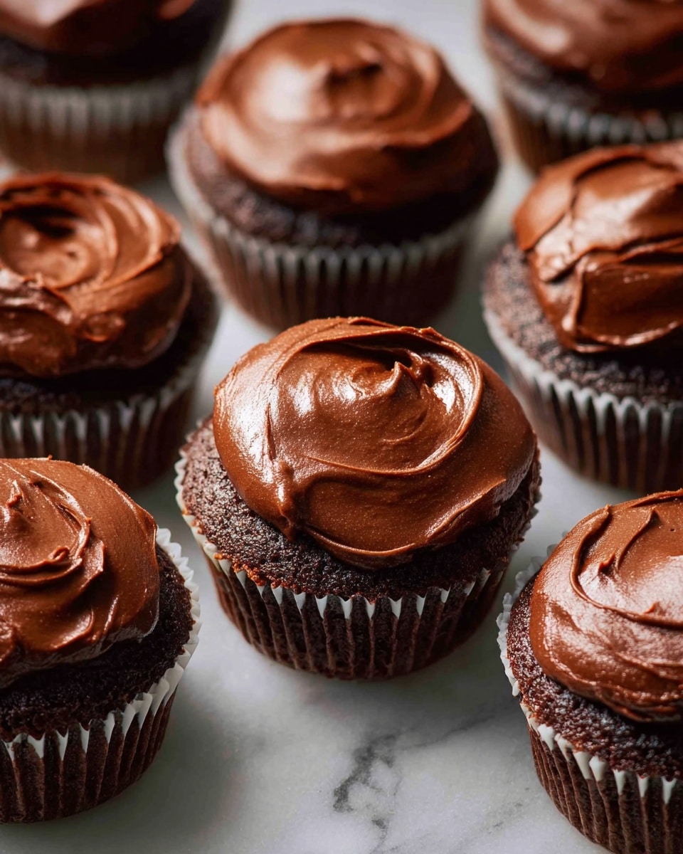 The image shows multiple chocolate cupcakes arranged close together on a white marbled surface. Each cupcake has two layers: a dark brown, moist cake base and a thick, glossy layer of smooth chocolate frosting spread generously on top. The frosting has visible swirls and soft peaks, giving a rich texture to the surface of each cupcake. The cupcakes are in white paper liners, arranged neatly with slight spacing between them. The lighting highlights the shine on the frosting, making it look fresh and creamy photo taken with an iphone --ar 4:5 --v 7