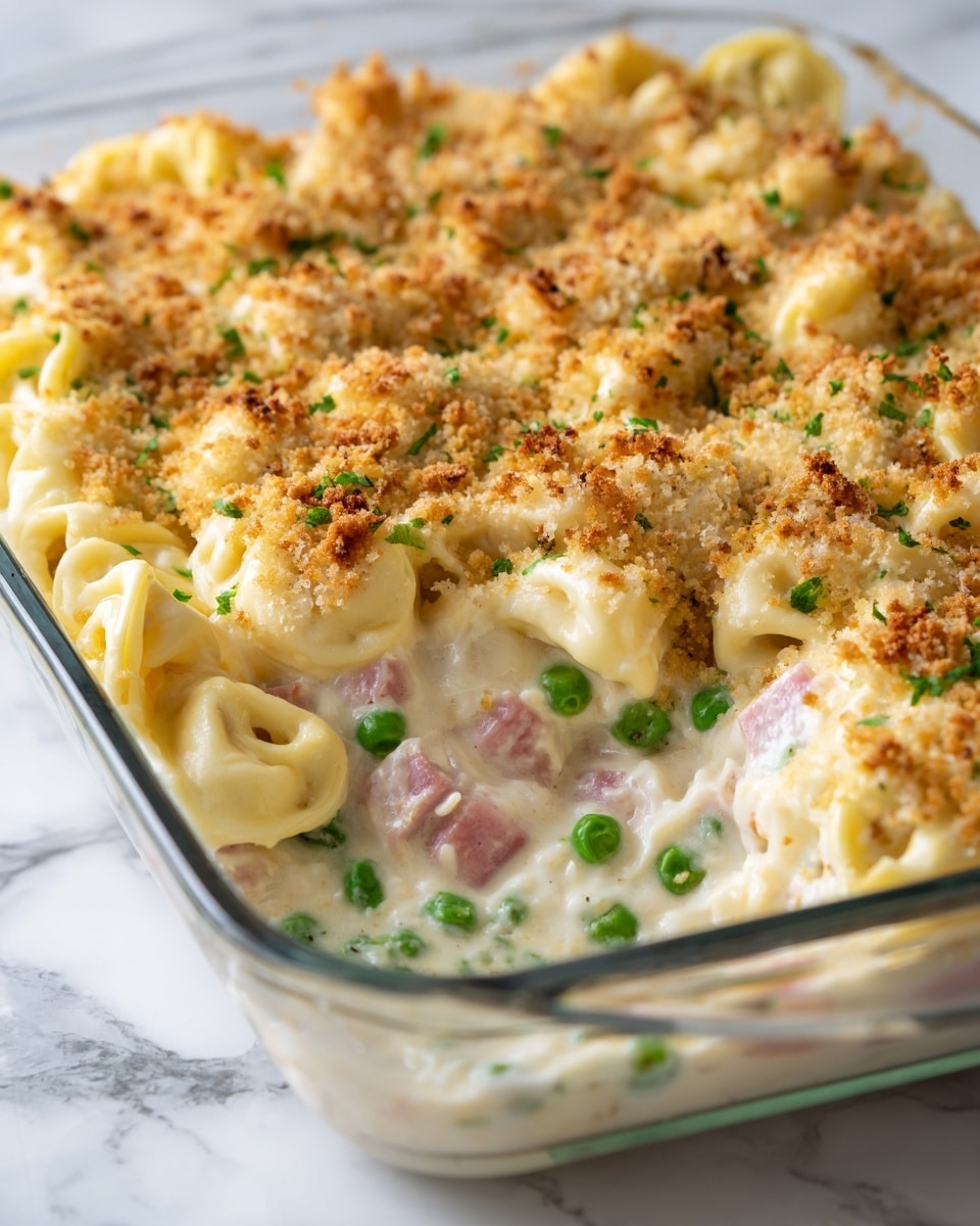 A close-up view of a glass baking dish filled with a creamy pasta casserole. The dish shows about three main layers: a base of tortellini pasta mixed with a creamy white sauce, scattered green peas, and small pieces of ham throughout. The top layer is sprinkled with a light brown crumb topping, adding texture. Part of the casserole has been scooped out, revealing the mix inside with soft, pale yellow pasta coated in the sauce, bright green peas, and pink ham cubes. The dish is set on a white marbled surface in the background, and photo taken with an iphone --ar 4:5 --v 7