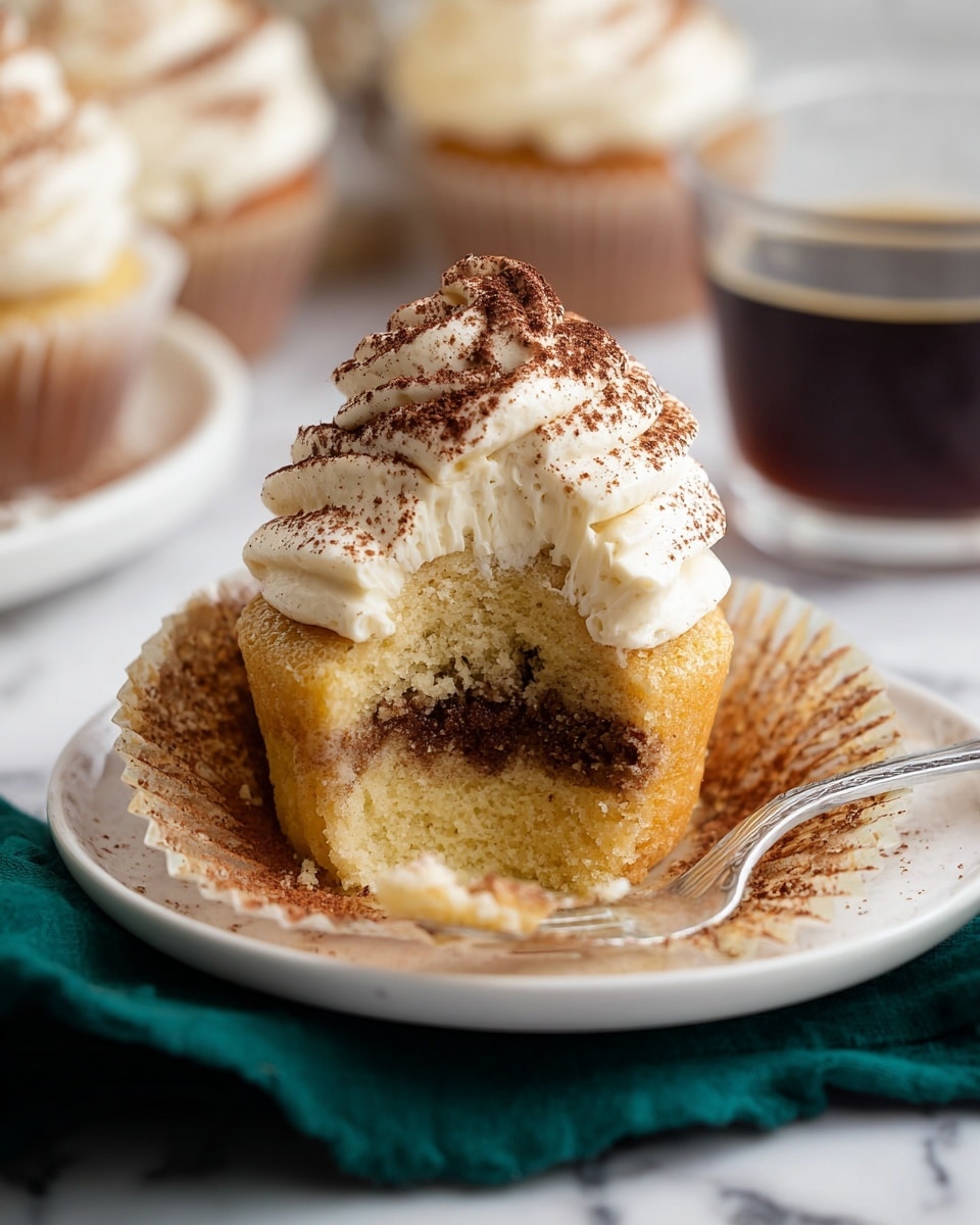 A partly eaten cupcake sits on a white plate with its paper wrapper peeled down, showing two layers: a light yellow bottom cake layer and a darker brown top layer. On top of the cupcake is a thick, creamy white frosting swirled high with a dusting of cocoa powder. In the background, there are blurred cupcakes and a clear cup with dark coffee. A silver fork with a bit of cupcake and frosting rests on the right side of the plate, and a dark green cloth is draped under the plate on a white marbled surface. photo taken with an iphone --ar 4:5 --v 7