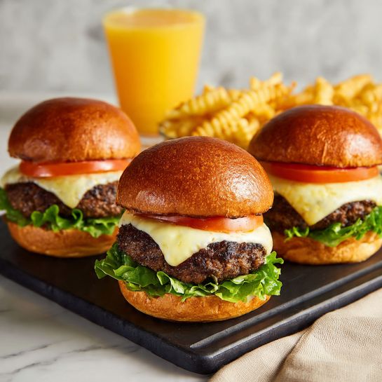 A black air fryer basket sits on a white marbled surface, containing four cooked hamburger patties. The patties are brown with a slightly shiny, cooked texture and round shapes with uneven edges. A metal spatula is lifting the top right patty, showing a flat, smooth silver surface and straight handle. The inside of the air fryer basket has a grid pattern that allows air circulation and small ridges along the sides. The top of the basket shows ventilation slots and a clear plastic window on the handle area. photo taken with an iphone --ar 4:5 --v 7