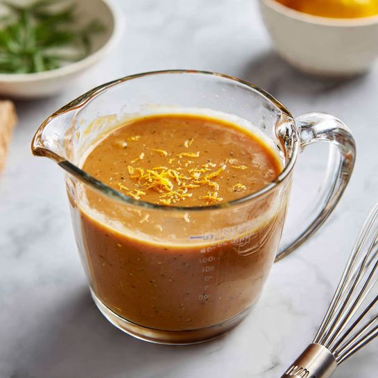 A clear glass measuring cup filled halfway with a thick, light brown sauce that has small bits and specks in it. The inside of the cup near the top is smeared with orange zest, adding a bright contrast to the sauce. The measuring cup has a handle on the right side and is placed on a white marbled surface. To the right of the cup is a silver whisk resting on the surface. In the blurred background, there's a white bowl with some green herbs inside, and a hint of yellow fruit is visible. Photo taken with an iphone --ar 4:5 --v 7