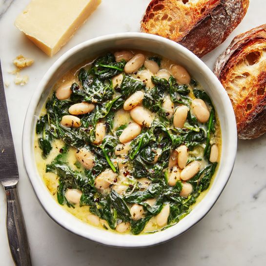 A white bowl filled with a creamy white bean and spinach dish, showing two main layers: light creamy white beans mixed with vibrant dark green spinach leaves spread evenly throughout. The bowl is placed on a white marbled surface, with two pieces of crusty, lightly golden brown bread positioned behind it, adding a rustic touch. A small wedge of pale yellow cheese is slightly visible in the top left corner, and a silver-handled knife rests to the left side. Photo taken with an iphone --ar 4:5 --v 7
