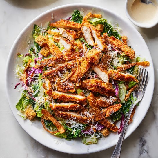 A white plate sits on a white marbled surface, holding a layered dish. The bottom layer is a mix of shredded green and purple cabbage, along with green kale leaves, creating a colorful, textured base. On top of this is a layer of light beige croutons scattered around, followed by a sprinkling of finely grated white cheese. The top layer consists of several slices of golden brown breaded chicken, each showing a clear white interior and a crispy dark golden crust. The chicken slices are neatly placed close together, covering most of the base layers. photo taken with an iphone --ar 4:5 --v 7