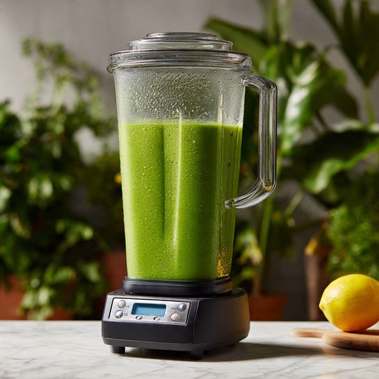 A close-up of a tall blender filled with a bright green smoothie, showing the thick, slightly textured liquid inside. The blender jar is clear plastic, with condensation on the inside near the top where the smoothie reaches, and a clear lid securely in place. The smoothie's vibrant green color stands out against the background of green plants and a yellow lemon on a wooden surface below. The blender sits on a black base with a digital display and several buttons. The whole scene is set on a white marbled surface with soft, natural lighting. photo taken with an iphone --ar 4:5 --v 7