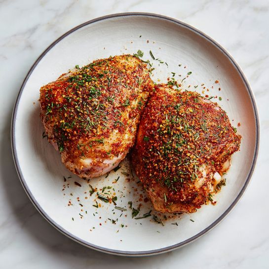 The image shows two pieces of raw chicken thighs lying flat side by side on a white plate with a thin gray rim. The chicken pieces are covered with a reddish-brown spice mix that looks coarse with visible bits of herbs and granules, evenly spread over the top layers of the meat. The plate rests on a white marbled surface with a clean and minimalistic background. photo taken with an iphone --ar 4:5 --v 7
