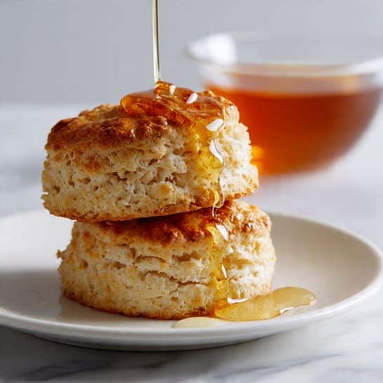 Two golden brown biscuit pieces are stacked on a white plate, resting on a white marbled surface. The top biscuit piece is fluffy and textured with a rough crust, while the bottom piece shows a soft, porous interior. Honey is drizzling down from above onto the top biscuit, creating shiny drops on its surface. In the background, a clear glass bowl filled with honey is partially visible, slightly out of focus. photo taken with an iphone --ar 4:5 --v 7