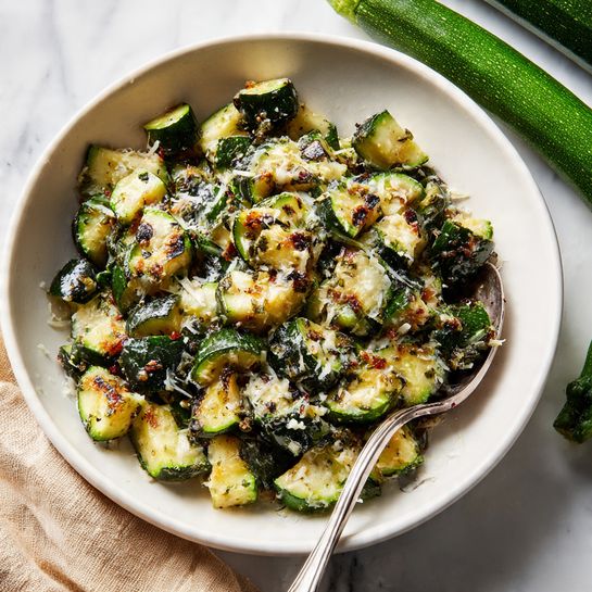 A white bowl filled with two layers of sautéed zucchini pieces, showing a mix of soft light green and darker green skin with light char marks and small bits of grated white cheese sprinkled on top; a silver spoon rests inside the bowl near the front, and two whole zucchinis are visible in the blurred white marbled background, along with a beige cloth on the left side. Photo taken with an iphone --ar 4:5 --v 7