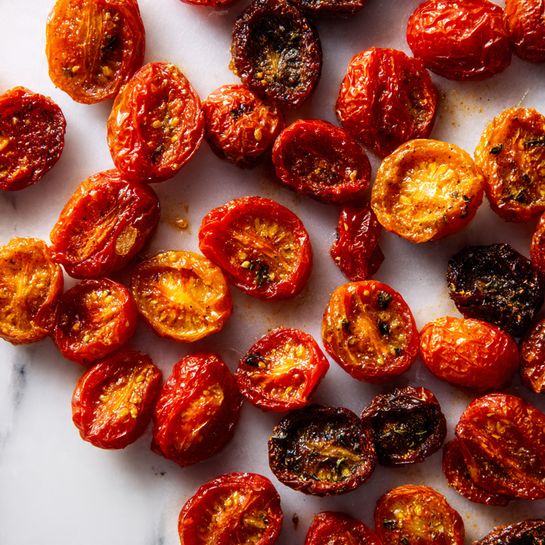 This image shows many roasted cherry tomatoes cut in half spread out on a white marbled surface. The tomatoes vary in color, mostly red and orange, with a few dark brownish ones. Each tomato half has a slightly wrinkled texture and some caramelized edges. The spread looks uneven but full, with tomatoes placed close to each other but not stacked. The white marbled background has slight marks from roasting and adds a clean, bright contrast to the colorful tomatoes. photo taken with an iphone --ar 4:5 --v 7