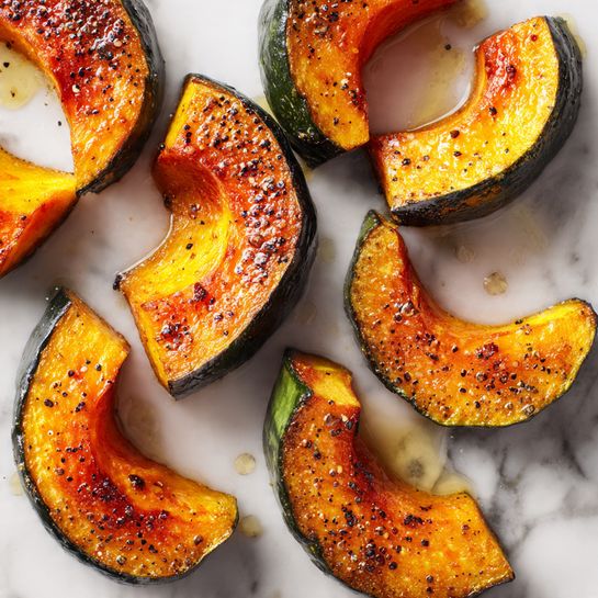 The image shows several pieces of roasted orange squash placed on a white marbled surface. Each piece has a thick slice with the dark green skin still visible on one side, while the inside is a warm orange color, browned and caramelized on top with a slightly rough texture. Small black pepper specks and glistening oil droplets are scattered around the squash, adding a shiny, moist appearance. The pieces vary in shape but all share a roasted, crispy edge look. photo taken with an iphone --ar 4:5 --v 7