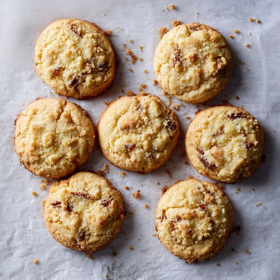 Six crumbly cookies with a light golden color sit on a sheet of parchment paper, arranged in two columns of three. Each cookie has a textured top layer with small crunchy bits and visible darker brown spots suggesting baked nuts or caramel pieces. The edges of the cookies are slightly more browned and uneven, creating a rustic look. Crumbs dot the parchment around the cookies, adding to the homemade feel. The background is a white marbled texture. Photo taken with an iphone --ar 4:5 --v 7