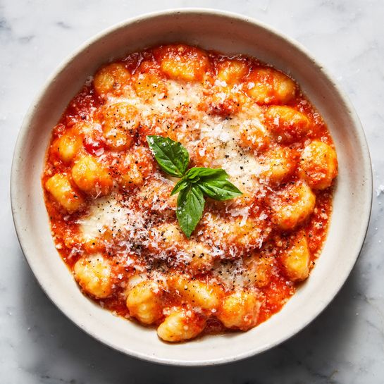 A white bowl filled with soft gnocchi covered in a thick, bright orange tomato sauce mixed with melted white cheese that has slightly browned on top. The gnocchi are small and rounded, with a bumpy texture visible through the sauce. There is a single green basil leaf placed near the center on top, and a sprinkle of grated cheese with some black pepper scattered over the dish. The bowl is sitting on a white marbled surface with some salt or cheese crumbs around it, giving a fresh and rustic feel. photo taken with an iphone --ar 4:5 --v 7