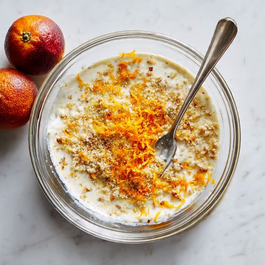 A clear glass bowl sits on a white marbled surface, filled with a mix that has a creamy white base topped with a layer of fine granules and bright orange zest sprinkled in the center. A metal fork with a long handle stands upright in the bowl, slightly pressing into the mixture. In the top left corner, two whole blood oranges with reddish-orange and brownish skins rest on the surface. Photo taken with an iphone --ar 4:5 --v 7