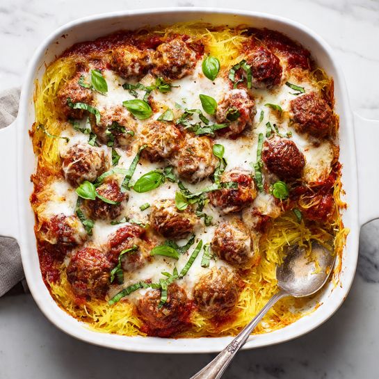 The image shows a white baking dish filled with a layered casserole. The bottom layer is golden-yellow spaghetti squash strands, soft and stringy. On top, there are thick red tomato sauce patches mixed with browned meatballs. Melted white cheese is spread unevenly over the meatballs and sauce, slightly browned in spots. Fresh green basil leaves are scattered on the top as garnish. A silver spoon rests inside the dish with a portion scooped out from one corner. The dish sits on a white marbled surface. Photo taken with an iphone --ar 4:5 --v 7