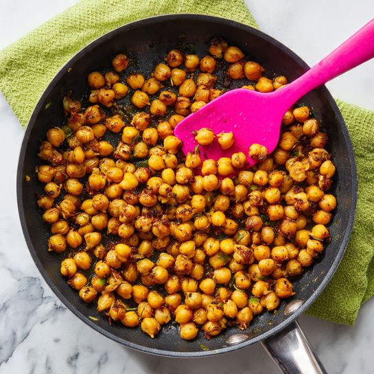 A black pan filled with a single layer of golden-brown cooked chickpeas, some showing slight toasting and light browning, mixed with small bits of green herbs or vegetables. A bright pink silicone spatula is partially buried in the chickpeas, resting near the center of the pan. The pan is placed on a green towel over a white marbled surface. Photo taken with an iphone --ar 4:5 --v 7