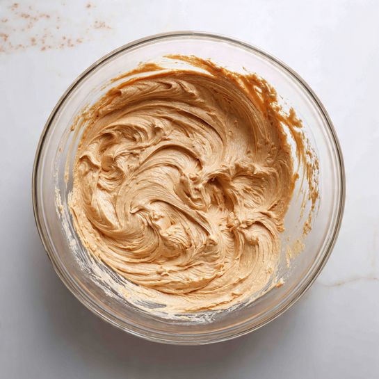 A clear glass bowl holds whipped light brown batter with visible texture and slight streaks in different shades of brown. The batter clings to the sides and bottom in swirled patterns, revealing the smooth glass underneath. The bowl rests on a white marbled surface with light beige speckles. There are no other items or colors in the scene, focusing fully on the creamy, swirled batter inside the bowl. photo taken with an iphone --ar 4:5 --v 7