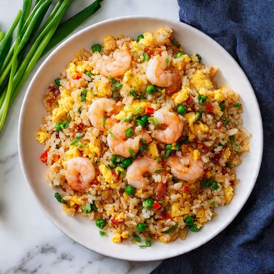 A deep white bowl filled with fried rice showing multiple layers: a base of golden brown cooked rice mixed with small bright green peas, orange carrot cubes, and red bell pepper pieces. On top, plump, pale pink shrimp are scattered with small fluffy yellow scrambled egg bits, all mixed with fresh green parsley leaves and chopped green onions. The bowl sits on a matching white plate on a white marbled surface, accompanied by light wooden chopsticks placed diagonally beside the plate. In the background, part of another white bowl with the same dish is slightly blurred along with a small bowl of green herbs and some green onions. Photo taken with an iphone --ar 4:5 --v 7