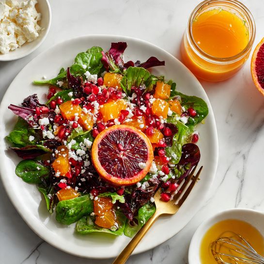 A white plate filled with a colorful salad showing different layers and textures. The bottom layer is mixed dark and light green leafy lettuce. On top of the leaves are small orange cubes, red pomegranate seeds, and irregular cuts of dark red blood orange pieces. White crumbled cheese is scattered across the salad. A bright slice of blood orange rests on the upper left edge of the plate. A gold fork is placed beside the salad on the right. The plate sits on a white marbled textured surface. Photo taken with an iphone --ar 4:5 --v 7