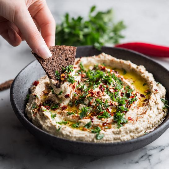 A dark grey bowl holds a creamy beige dip with a slightly rough texture. The dip is topped with sprinklings of finely chopped green herbs and bright red chili flakes. There is a drizzle of golden oil, pooling gently in a shallow swirl on top. A woman's hand is seen dipping a triangular piece of dark brown bread into the dip. In the background, some fresh herbs and a red chili lie on a white marbled surface. photo taken with an iphone --ar 4:5 --v 7