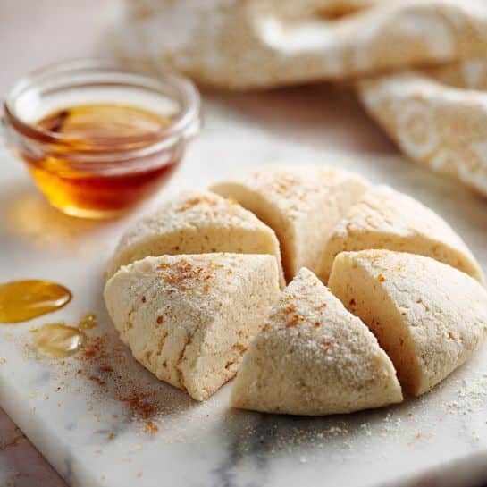 A round dough piece rests on a white marbled surface, divided into six triangular parts, showing a soft and slightly crumbly texture with some flour dusted around it. To the left, there is a small clear glass bowl with golden honey and a drop of honey on the white marbled surface nearby. The background is softly blurred with a hint of a decorative cloth. photo taken with an iphone --ar 4:5 --v 7