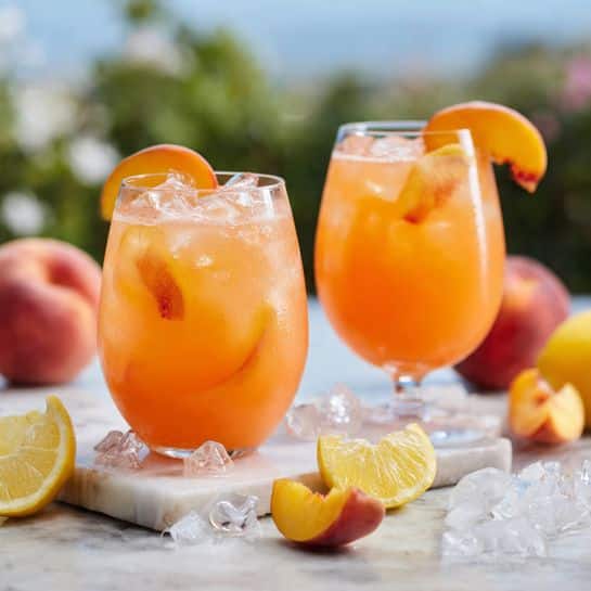 A close-up top view of a clear round glass filled with an orange-colored drink with ice cubes. Inside the drink, there are two peach slices placed near the top. The glass sits on a white marbled surface scattered with ice cubes, with blurred peach halves and other glasses partially visible around the edges. The drink has a light, refreshing texture with condensation on the glass. photo taken with an iphone --ar 4:5 --v 7