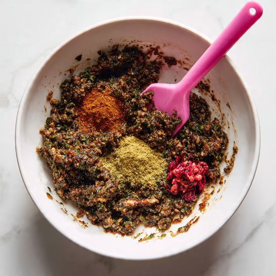 A white bowl filled with a grainy, mixed paste that is dark brown with green herb bits spread throughout. On top of the paste are several piles of dry spices in bright colors: a rusty red powder, a yellow-green powder, and a small spot of bright red paste. A pink spatula is partially submerged on the right side of the bowl, mixing the contents. The bowl rests on a white marbled surface. photo taken with an iphone --ar 4:5 --v 7
