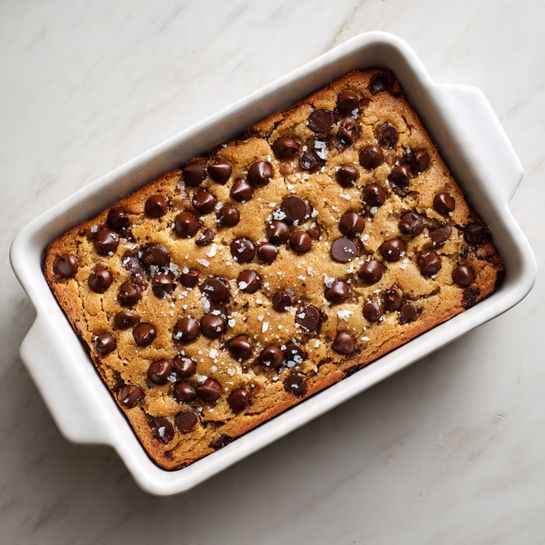 A baked chocolate chip cookie bar is shown inside a white rectangular ceramic dish. The cookie bar has one thick layer with a golden brown baked surface, dotted with evenly spaced dark brown chocolate chips, some partially melted. There are also small flakes of white sea salt sprinkled on top, creating a contrast against the golden cookie base and chocolate chips. The dish is placed on a white marbled surface. photo taken with an iphone --ar 4:5 --v 7
