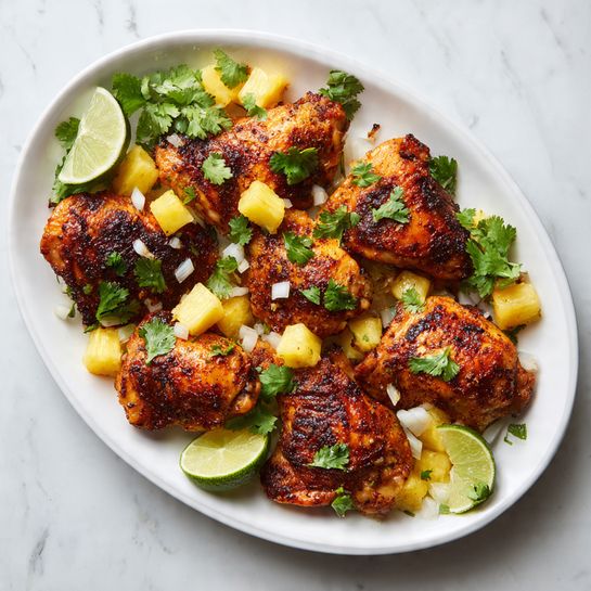 A white oval plate filled with about nine golden-brown roasted chicken pieces, some with a slightly dark, crispy char. On top of the chicken is a fresh mix of small yellow pineapple chunks, white onion pieces, and green cilantro leaves spread evenly. Two lime halves, one slightly squeezed, rest on the edge of the plate. The plate sits on a white marbled surface. photo taken with an iphone --ar 4:5 --v 7