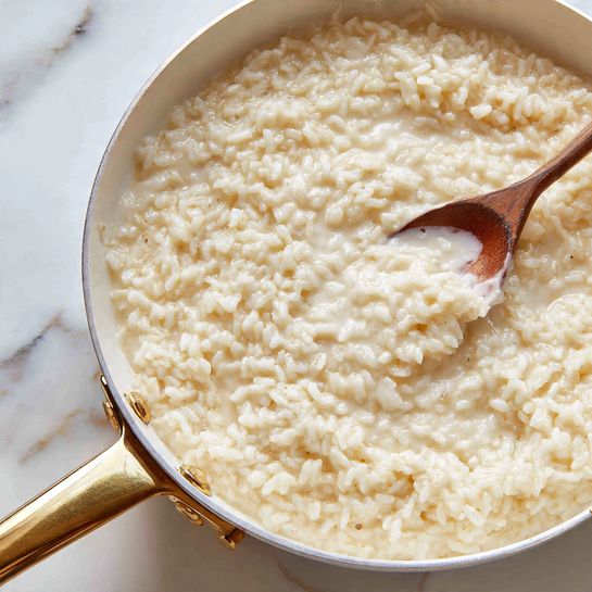A close-up view of a creamy rice dish served in a white pan with a small part of its gold handle visible at the bottom. The rice appears soft and sticky, with a wet, shiny texture showing each grain coated in a thick, pale yellow sauce. The background is a white marbled surface, adding a clean and simple look to the image. photo taken with an iphone --ar 4:5 --v 7