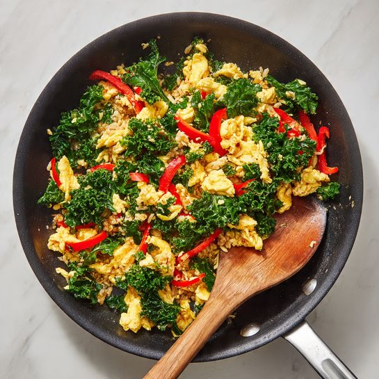 A black frying pan filled with a colorful stir-fry mixture. The dish has four main layers: fluffy yellow scrambled eggs scattered across the stir-fry, bright green kale leaves cooked to a soft texture, thin strips of red bell pepper adding a pop of color, and small pieces of light brown cooked rice that blend well with the other ingredients. A wooden spatula is resting on the side inside the pan, partially covered by the food. The pan sits on a white marbled surface. Photo taken with an iphone --ar 4:5 --v 7