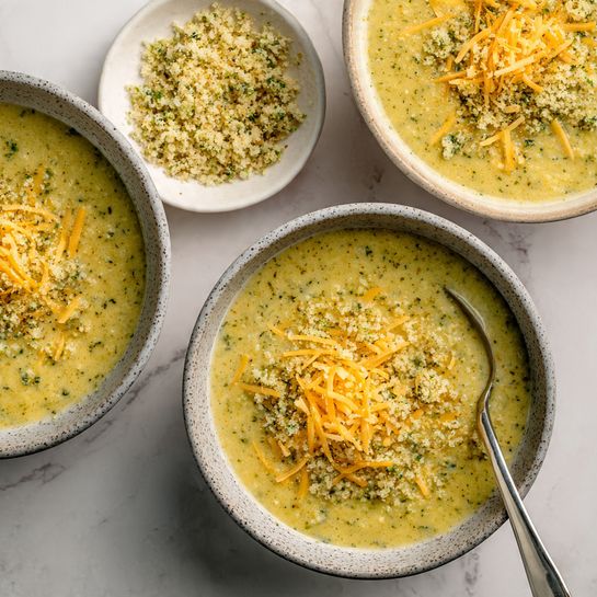 The image shows a thick yellow-green soup served in three round bowls with a stone-like texture on the outside and white inside, placed on a white marbled surface. Each bowl of soup has a thick, smooth texture with small green leafy pieces mixed in. The middle bowl has a spoon standing upright and is topped with shredded orange cheese and a crumbly mixture made of breadcrumbs and herbs, adding texture and color contrast. Next to the bowls, there are two silver spoons and a small white bowl filled with more of the crumbly herb topping. The scene is bright and clean, with soft natural light highlighting the soup’s creamy texture and fresh garnishes. photo taken with an iphone --ar 4:5 --v 7