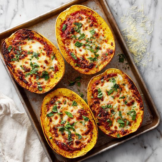 The image shows four halves of roasted spaghetti squash on a baking tray. Each squash half has a bright orange base layer with a browned, bubbly cheese topping that has patches of golden and dark brown textures. There are spots of red sauce scattered over the cheese, and fresh green herb leaves sprinkled on top. The tray rests on a white marbled surface with some cheese flakes and herb leaves scattered around, and a white cloth is partially visible on the lower left side. photo taken with an iphone --ar 4:5 --v 7