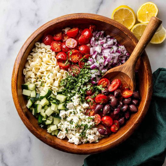 A close-up view of a white bowl filled with a colorful orzo salad, showing three layers: the base layer is light yellow orzo pasta with a soft texture, mixed evenly with small pieces of bright green cucumber and finely chopped dark green herbs; the middle layer includes halved bright red cherry tomatoes and slices of deep purple black olives scattered around; on top, there are crumbled white feta cheese and small pieces of chopped purple-red onion adding contrast. Two wooden spoons are partly visible inside the bowl, lifting some salad. The background is a white marbled texture. photo taken with an iphone --ar 4:5 --v 7