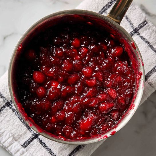 A close-up of a silver metal pot filled with a chunky, deep red cranberry sauce made of whole and crushed cranberries. The sauce has a glossy, slightly thick texture with visible pieces of softened fruit. The pot is resting on a white cloth with black grid lines, all placed on a white marbled surface. The inside edge of the pot is stained with splashes of the red sauce, showing freshness and recent preparation photo taken with an iphone --ar 4:5 --v 7