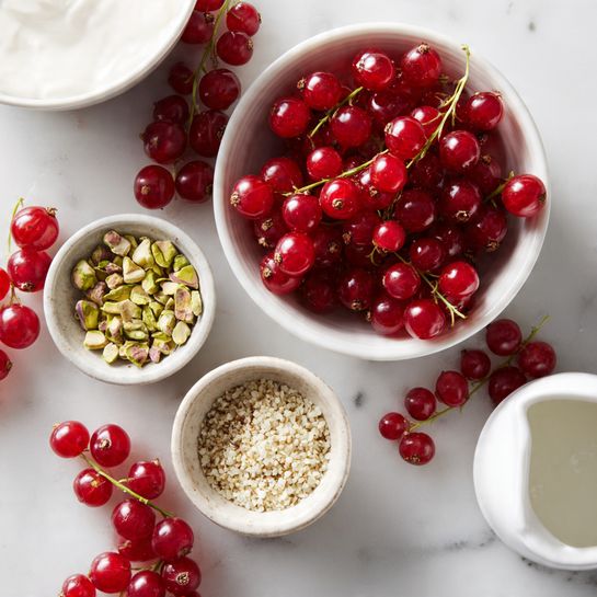 A white marbled surface holds several white bowls and clusters of bright red currants scattered around; one small white bowl contains light green and beige chopped pistachios, another small white bowl is filled with a small mound of pale hemp seeds with a slightly rough texture, and a white pitcher at the bottom right corner has a bit of smooth white liquid inside. Part of a larger white bowl with white curd is visible in the top left corner. The red currants are shiny and round, some still on their thin stems. Photo taken with an iphone --ar 4:5 --v 7