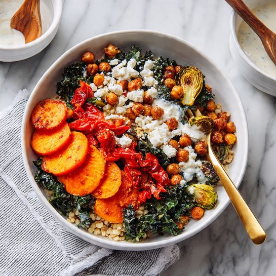 A white bowl filled with a colorful mix of roasted vegetables and toppings sits on a white marbled surface with a white cloth underneath. Starting from the top right, there are five thick, round slices of orange sweet potato with a slightly browned texture and sprinkled with seeds and salt. To the left of the sweet potatoes is a pile of roasted Brussels sprouts, browned and slightly charred on the edges, showing green and golden hues. Below the Brussels sprouts, dark red roasted bell pepper pieces add a pop of color. In the center, a thick layer of golden-brown roasted chickpeas is scattered generously, some overlapping the vegetables. Small crumbles of white cheese are sprinkled across the chickpeas and Brussels sprouts, adding contrast. Behind the bowl, a wooden spoon dips into a cup with light beige sauce, all set on the white marbled surface. Photo taken with an iphone --ar 4:5 --v 7