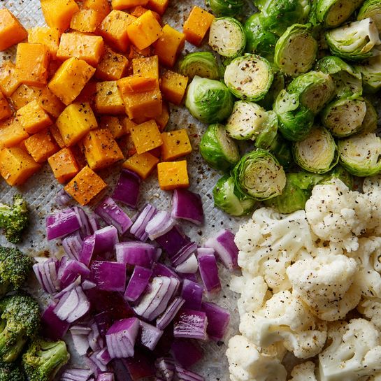 The image shows a close-up of six different kinds of chopped vegetables spread out on a baking tray. At the top left, there are orange cubes of squash with a slightly shiny texture. To the right of the squash, there are green Brussels sprouts, cut into halves showing layers of light green leaves with some black pepper sprinkled on top. Below the Brussels sprouts, purple and white pieces of diced onion form a mix of smooth and shiny cubes. At the bottom right, pieces of bright red bell pepper are scattered, showing shiny and smooth skin with some seeds visible. In the bottom left corner, small green broccoli florets with a rough texture are placed, and above them, white cauliflower florets showing their rough, bumpy texture. All vegetables have a light coating of oil and specks of black pepper. The background is a textured baking tray with a white marbled surface visible. Photo taken with an iphone --ar 4:5 --v 7