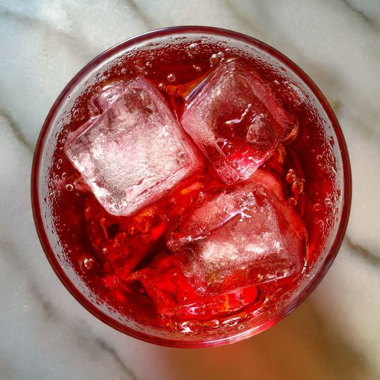 A close-up top view of a glass filled with a bright red drink, showing two large clear ice cubes and crushed ice, with droplets of condensation on the glass edges. The glass sits on a white marbled surface, with the rich red liquid contrasting against the subtle pale background. photo taken with an iphone --ar 4:5 --v 7