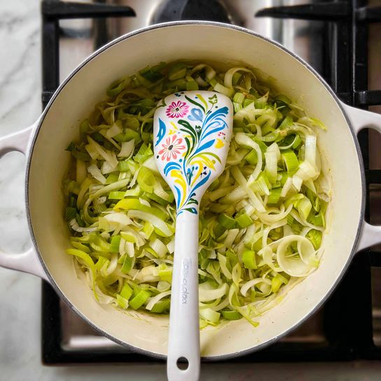 The image shows a large white pot filled with many thin, curly, light green and bright green sliced leeks. A white spatula with blue, yellow, and pink flower designs is placed upright in the middle of the leeks, stirring or resting. The pot is sitting on a black stove, and steam rises gently, showing the leeks are cooking. The white marbled surface is just barely visible around the edges. photo taken with an iphone --ar 4:5 --v 7